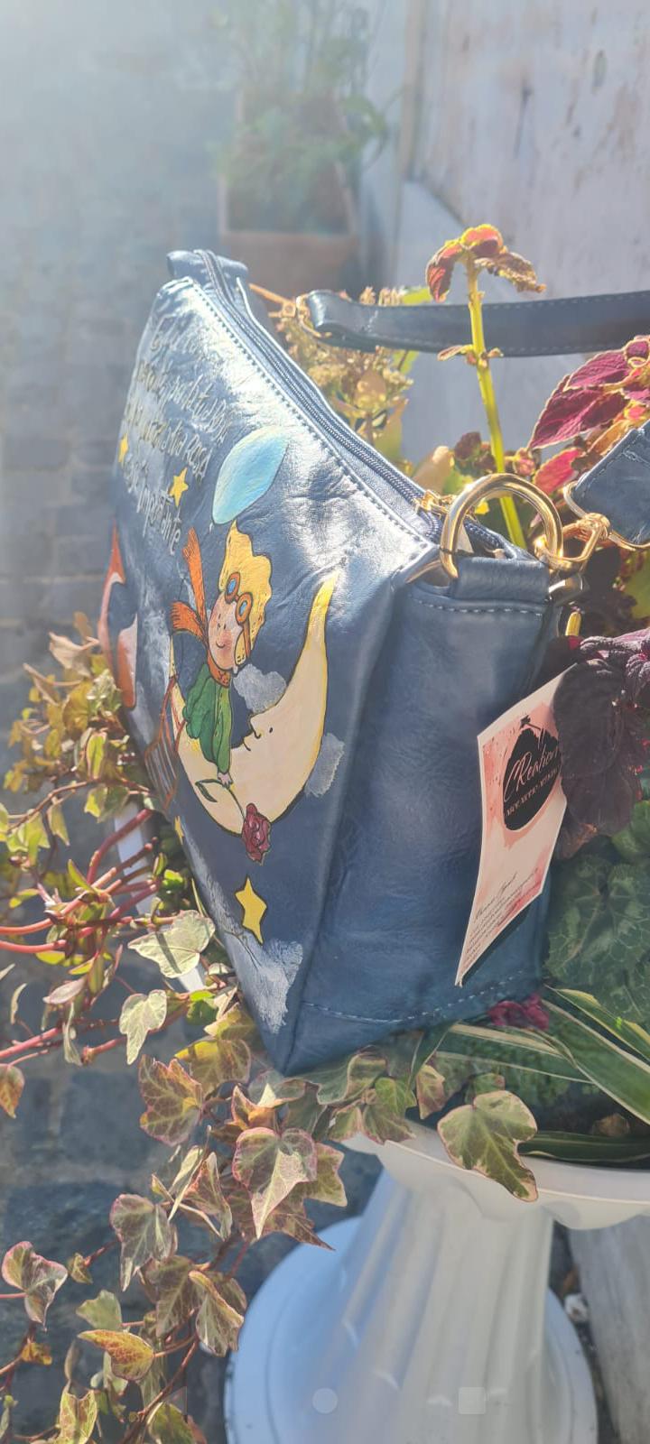 Borsa Dipinta a Mano - Il Piccolo Principe | Accessorio Unico e Magico Porta con te un pezzo di magia e saggezza con questa borsa dipinta a mano, che celebra il Piccolo Principe, il celebre personaggio creato da Antoine de Saint-Exupéry. Ogni borsa è realizzata con amore, utilizzando colori acrilici di alta qualità, per un design unico che racconta una storia di avventura, sogni e riflessioni. 🧩 *Coordinabile su richiesta:* È possibile ordinare *un portafoglio o un giacchetto perfettamente abbinato* con lo