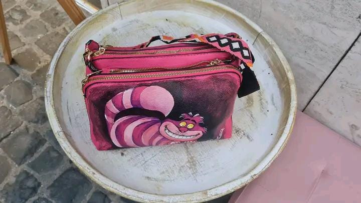 "Borsa Stregatto Alice nel Paese delle Meraviglie in Ecopelle | Dipinta a Mano | Lavabile fino a 40°C" 🧩 *Coordinabile su richiesta:* È possibile ordinare *un portafoglio o un giacchetto perfettamente abbinato* con lo stesso stile e soggetto ! Descrizione: Questa borsa unica nel suo genere è ispirata al famoso Stregatto di "Alice nel Paese delle Meraviglie".