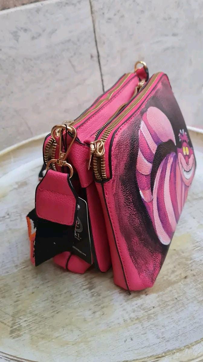 "Borsa Stregatto Alice nel Paese delle Meraviglie in Ecopelle | Dipinta a Mano | Lavabile fino a 40°C" 🧩 *Coordinabile su richiesta:* È possibile ordinare *un portafoglio o un giacchetto perfettamente abbinato* con lo stesso stile e soggetto ! Descrizione: Questa borsa unica nel suo genere è ispirata al famoso Stregatto di "Alice nel Paese delle Meraviglie".