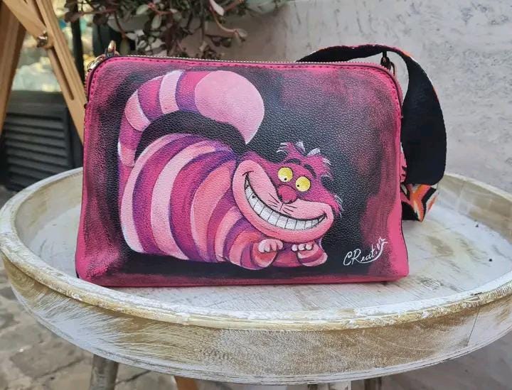 "Borsa Stregatto Alice nel Paese delle Meraviglie in Ecopelle | Dipinta a Mano | Lavabile fino a 40°C" 🧩 *Coordinabile su richiesta:* È possibile ordinare *un portafoglio o un giacchetto perfettamente abbinato* con lo stesso stile e soggetto ! Descrizione: Questa borsa unica nel suo genere è ispirata al famoso Stregatto di "Alice nel Paese delle Meraviglie".