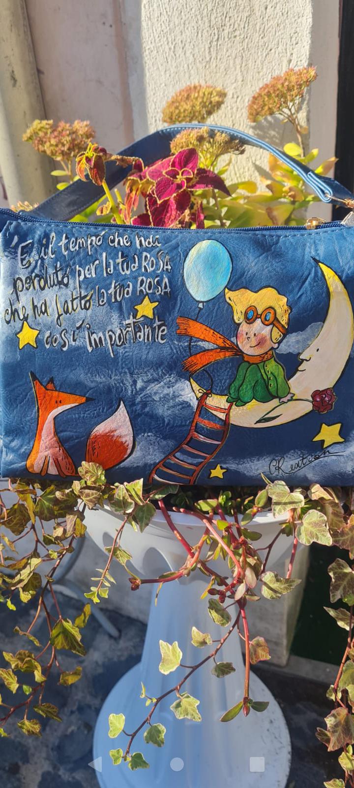 Borsa Dipinta a Mano - Il Piccolo Principe | Accessorio Unico e Magico Porta con te un pezzo di magia e saggezza con questa borsa dipinta a mano, che celebra il Piccolo Principe, il celebre personaggio creato da Antoine de Saint-Exupéry. Ogni borsa è realizzata con amore, utilizzando colori acrilici di alta qualità, per un design unico che racconta una storia di avventura, sogni e riflessioni. 🧩 *Coordinabile su richiesta:* È possibile ordinare *un portafoglio o un giacchetto perfettamente abbinato* con lo
