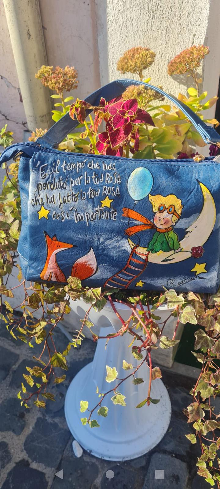 Borsa Dipinta a Mano - Il Piccolo Principe | Accessorio Unico e Magico Porta con te un pezzo di magia e saggezza con questa borsa dipinta a mano, che celebra il Piccolo Principe, il celebre personaggio creato da Antoine de Saint-Exupéry. Ogni borsa è realizzata con amore, utilizzando colori acrilici di alta qualità, per un design unico che racconta una storia di avventura, sogni e riflessioni. 🧩 *Coordinabile su richiesta:* È possibile ordinare *un portafoglio o un giacchetto perfettamente abbinato* con lo