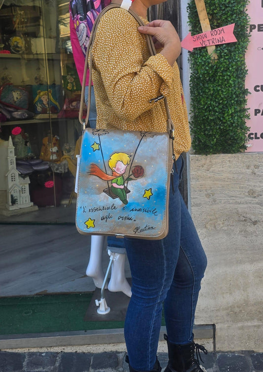 Borsa dipinta a mano – Il Piccolo Principe con frase iconica – Pelle/Ecopelle di alta qualità *Descrizione:* Lasciati ispirare dalla magia senza tempo de *Il Piccolo Principe* con questa borsa capiente, dipinta interamente a mano. Il soggetto principale è l’iconico protagonista del celebre racconto, accompagnato da una frase simbolica che emoziona grandi e piccini. Ogni borsa è realizzata su *pelle o ecopelle di alta qualità*, resistente e piacevole al tatto, e dipinta con *colori acrilici professionali* ad